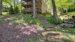 Eine zweistöckige Holzhütte mit einem Balkon, umgeben von Bäumen und einer Steintreppe, die zu ihr hinaufführt.