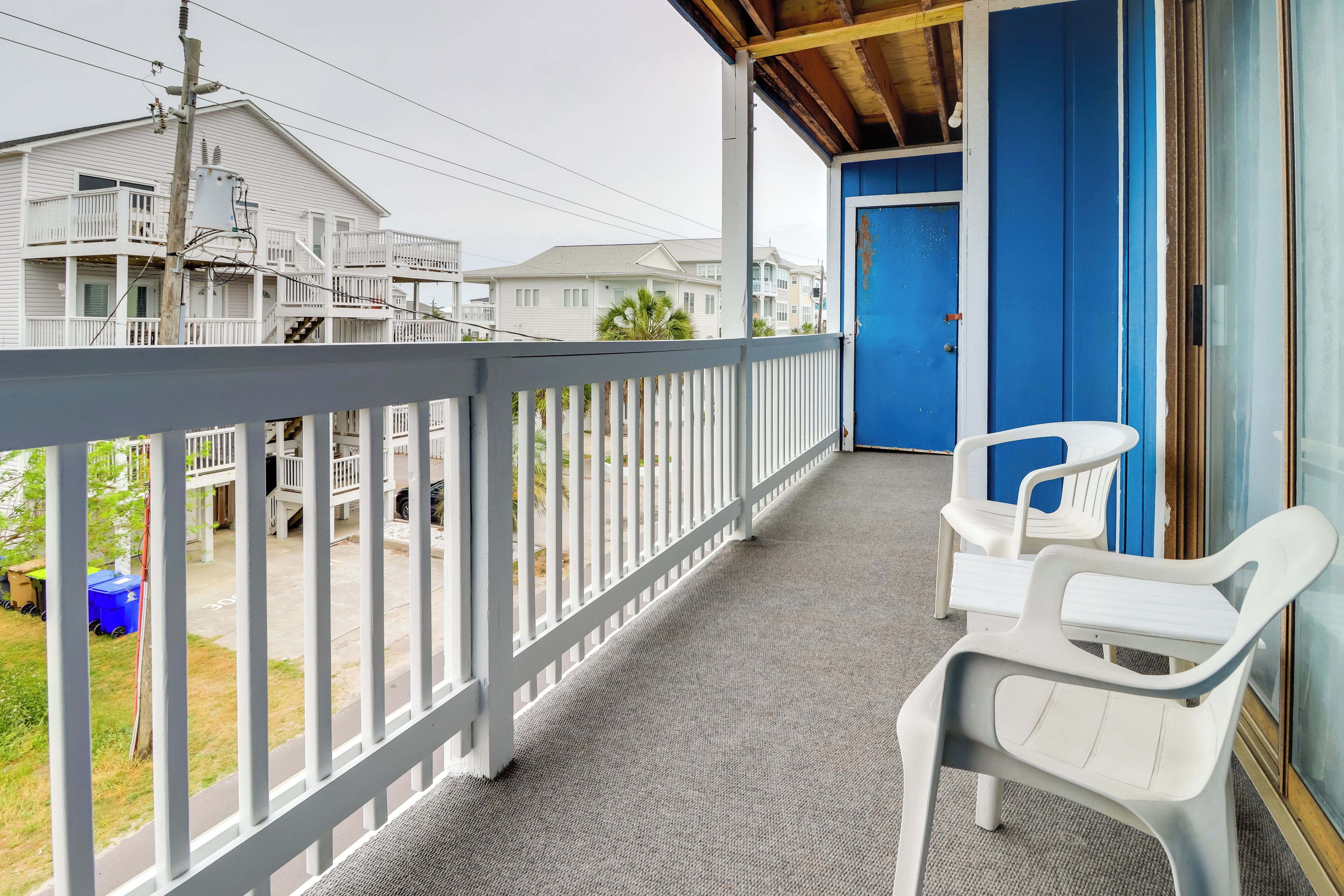Un balcon abrité, avec des chaises en plastique blanches, une porte bleue et une vue sur les maisons voisines.