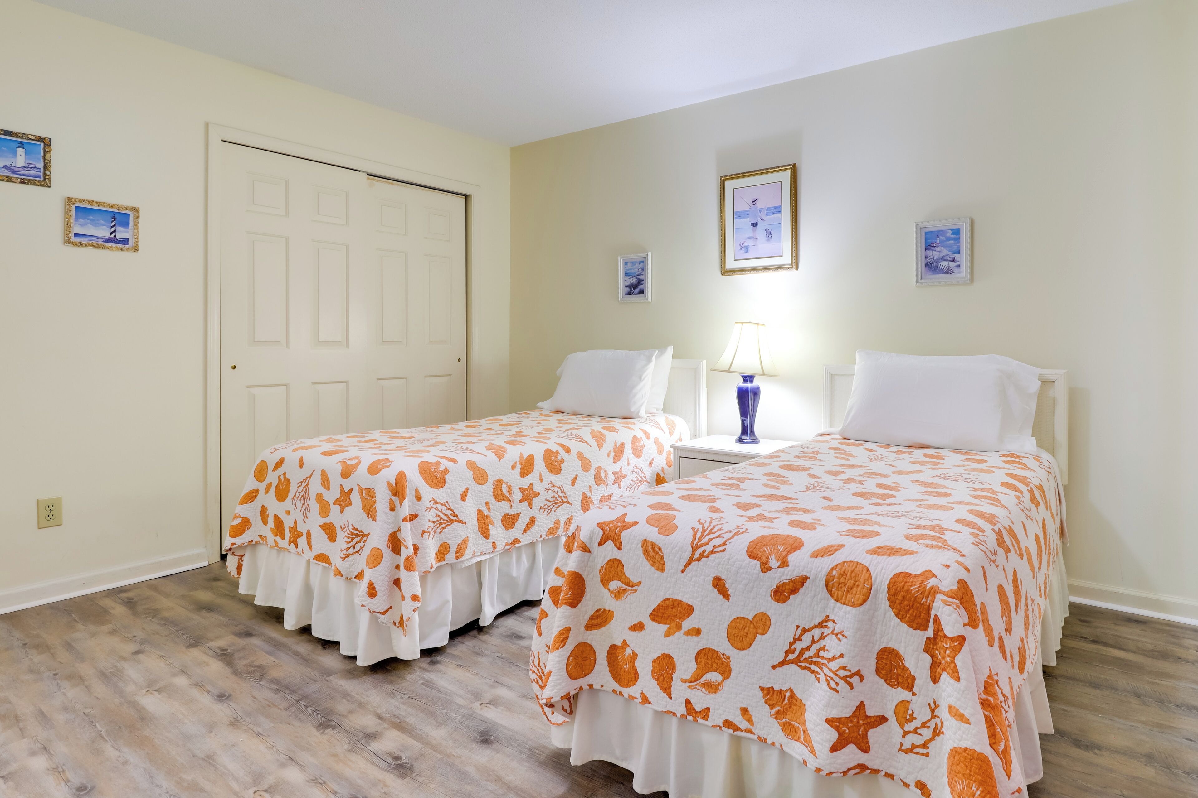Una habitación con dos camas, cada una con colcha blanca y naranja estampada, una puerta blanca y una mesita con lámpara.