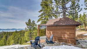 Una cabaña de madera con terraza de piedra, dos sillas Adirondack y vistas a un bosque y montañas.