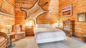 Un dormitorio de estilo cabaña de madera con una cama grande, una silla, una mesita, una estantería y un trozo de piel de animal colgado en la pared.