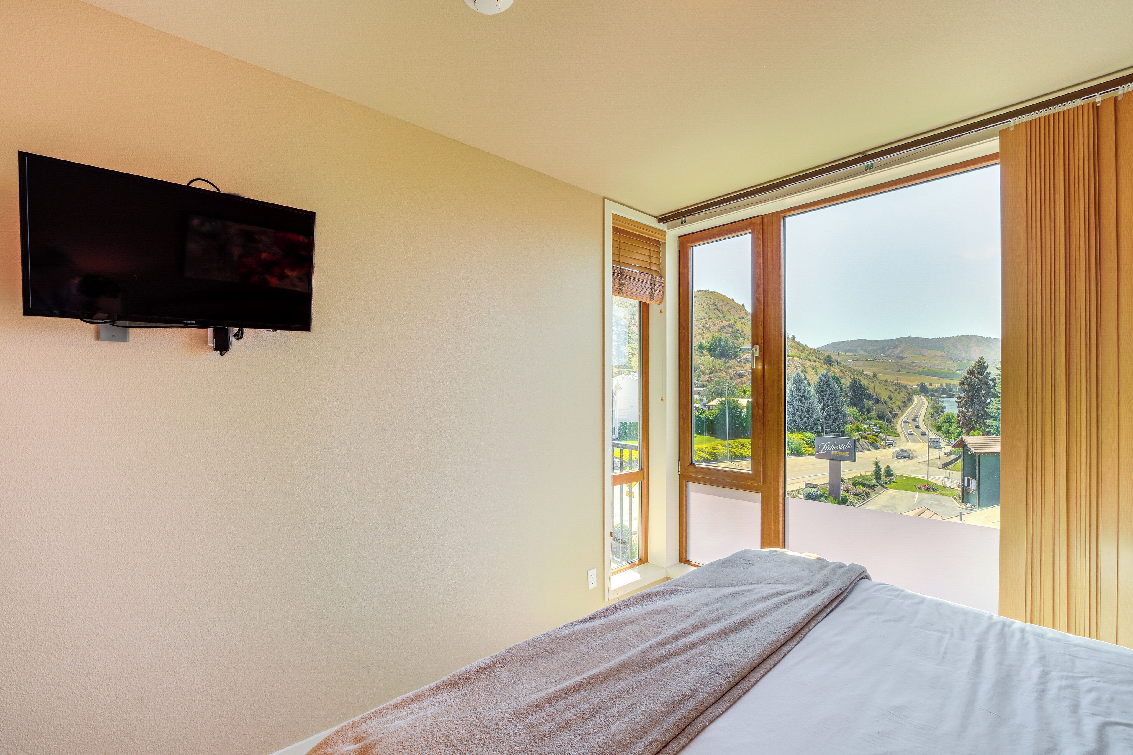 Habitació d'hotel amb un llit, televisió penjada a la paret i una finestra gran amb vista a un paisatge amb turons.