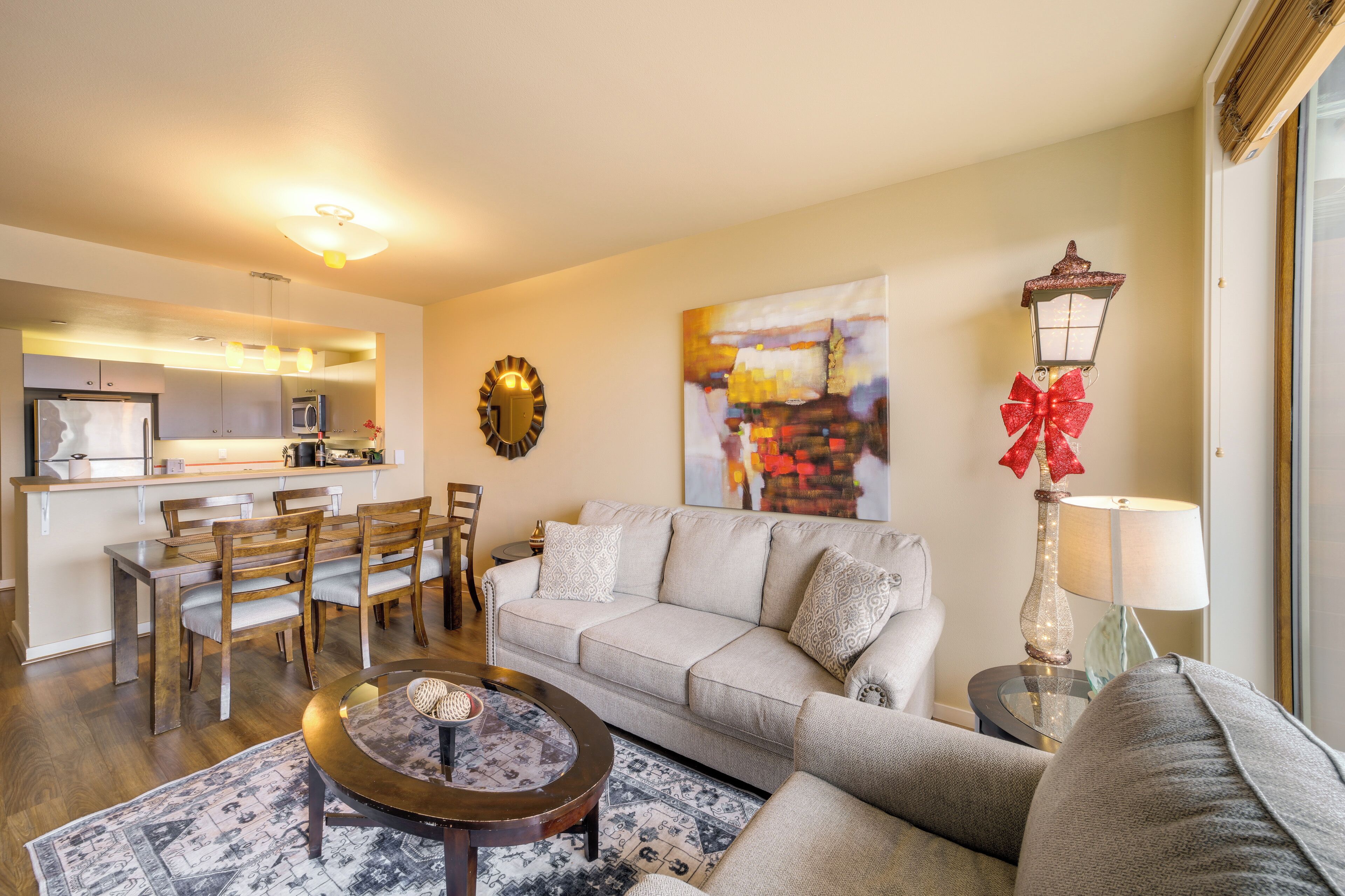 A living room with a sofa, coffee table, and dining area. There is a kitchen in the background with a refrigerator and microwave.