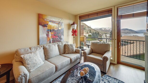 Un salon comprenant un canapé, un fauteuil et une table basse. Un tableau est accroché au mur, et on aperçoit un balcon avec une balustrade.