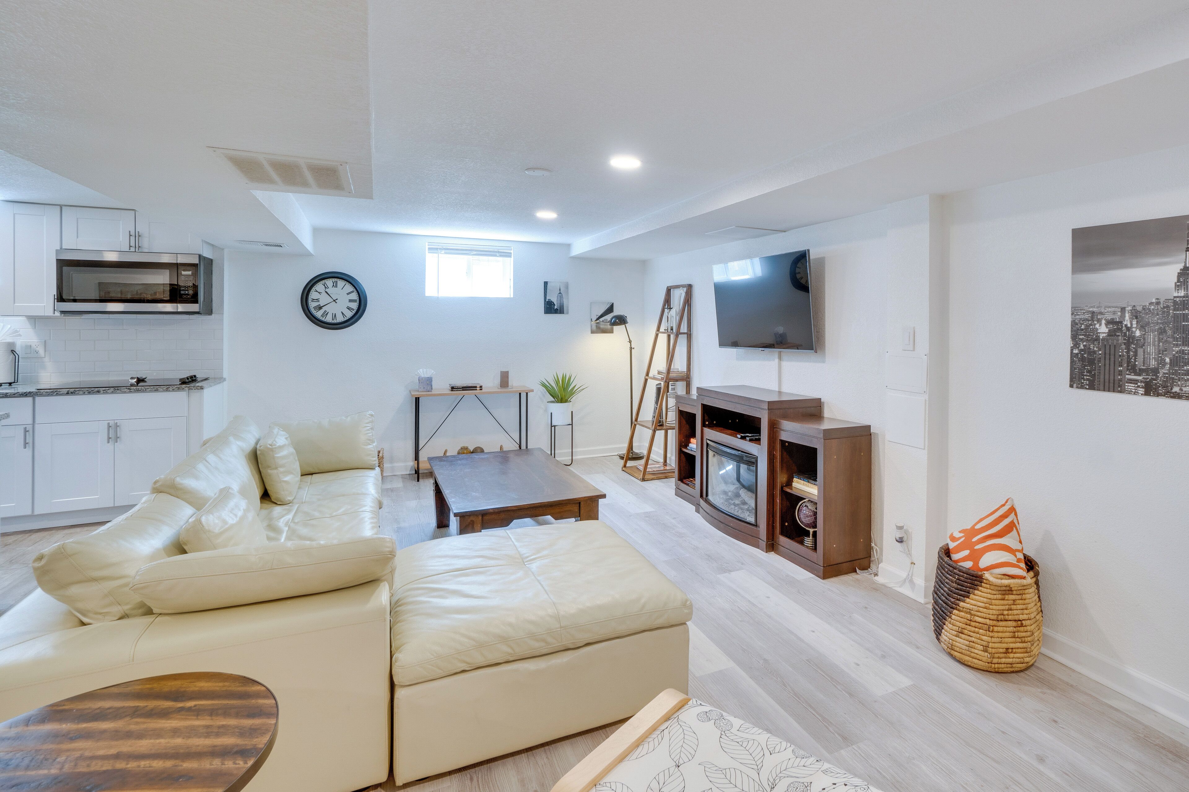 Ein modernes Wohnzimmer mit einer Couch, einem Couchtisch und einem Kamin. Es gibt eine Küchenzeile mit einer Mikrowelle und Schränken. Ein Fernseher ist an der Wand montiert, und eine Uhr ist sichtbar.