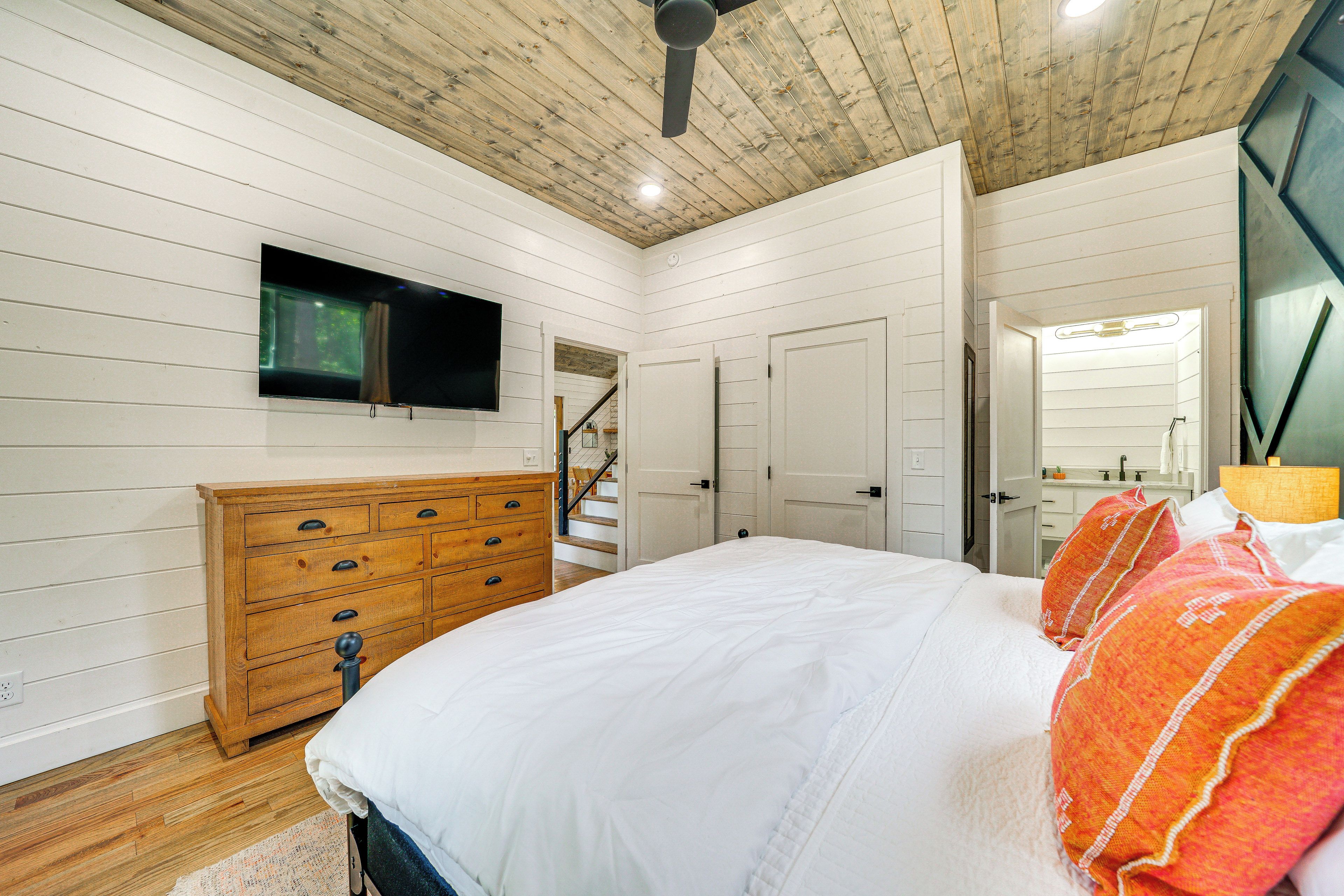 A bedroom with a wooden ceiling, a bed with white bedding, a wooden dresser, a television on the wall, and a staircase leading to another room.