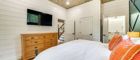 A bedroom with a wooden ceiling, a bed with white bedding, a wooden dresser, a television on the wall, and a staircase leading to another room.