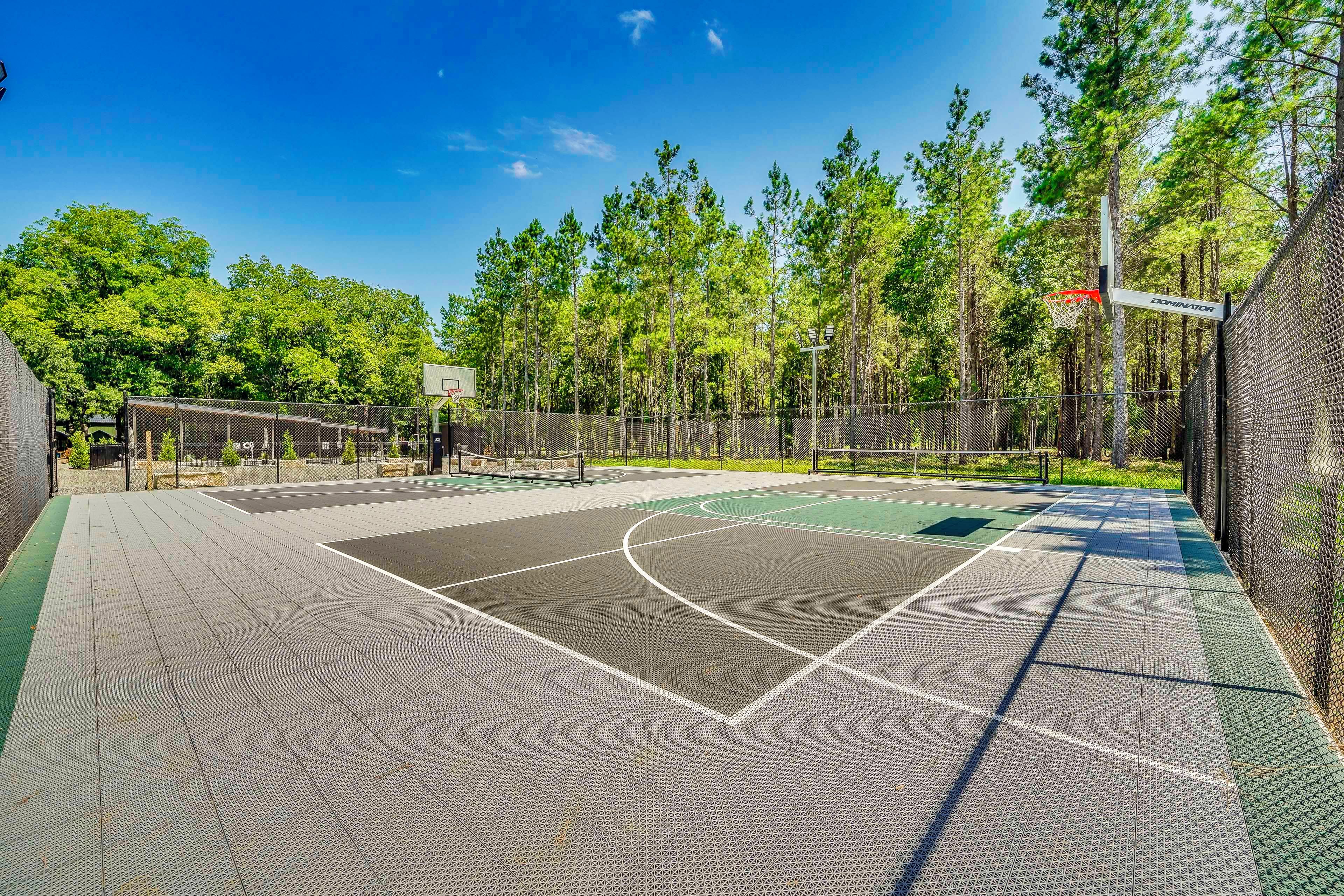 An outdoor basketball court with a chain-link fence, trees, and a clear sky.