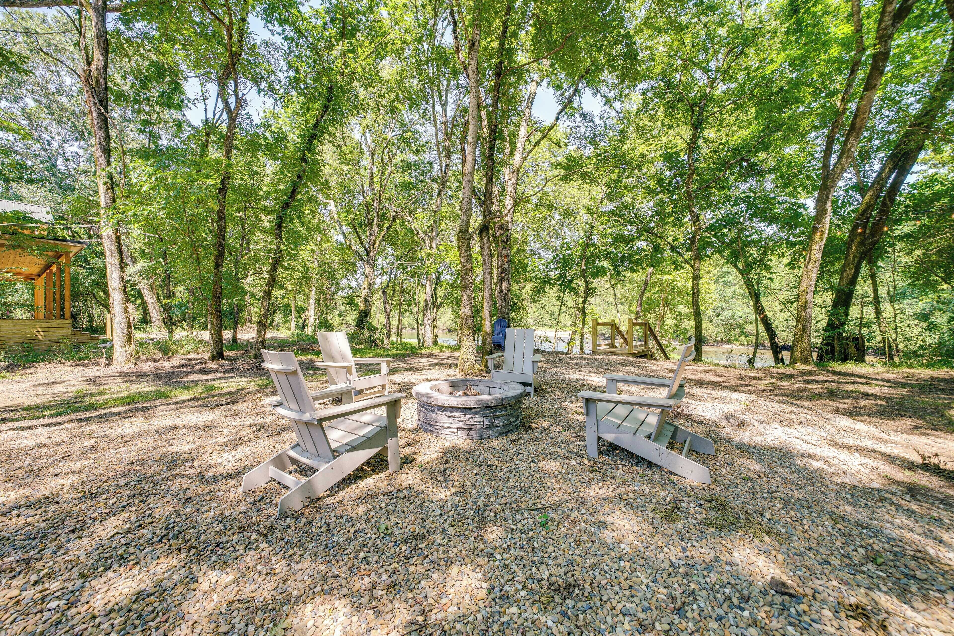 A forested area with a fire pit and Adirondack chairs.