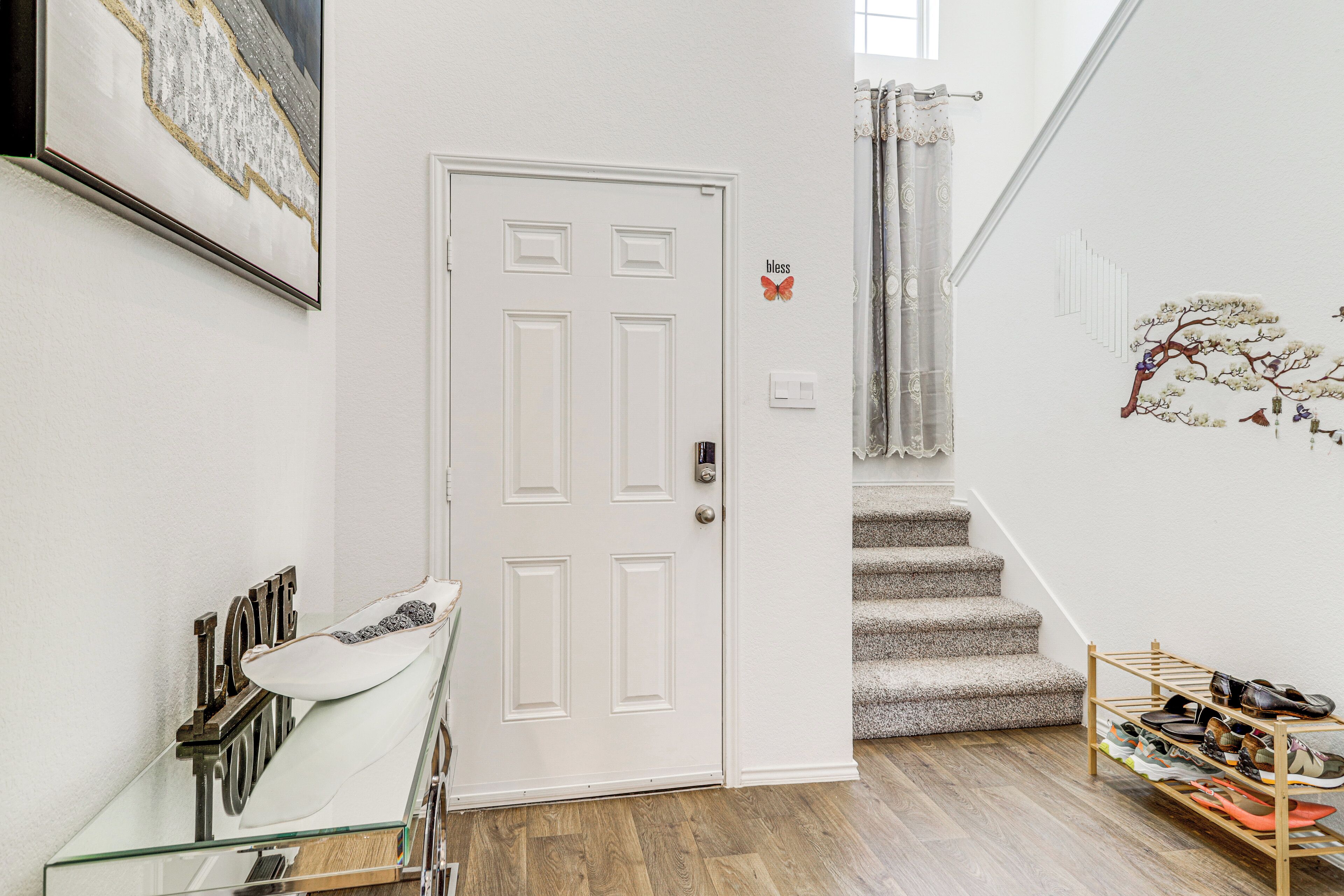 A staircase with a shoe rack at the bottom, a white door with a butterfly decoration, and a wall with a 'bless' sign.