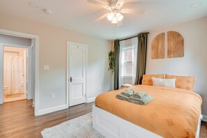 Une chambre à coucher comprenant un lit, un ventilateur de plafond, une fenêtre avec des rideaux et une porte donnant sur une autre pièce.