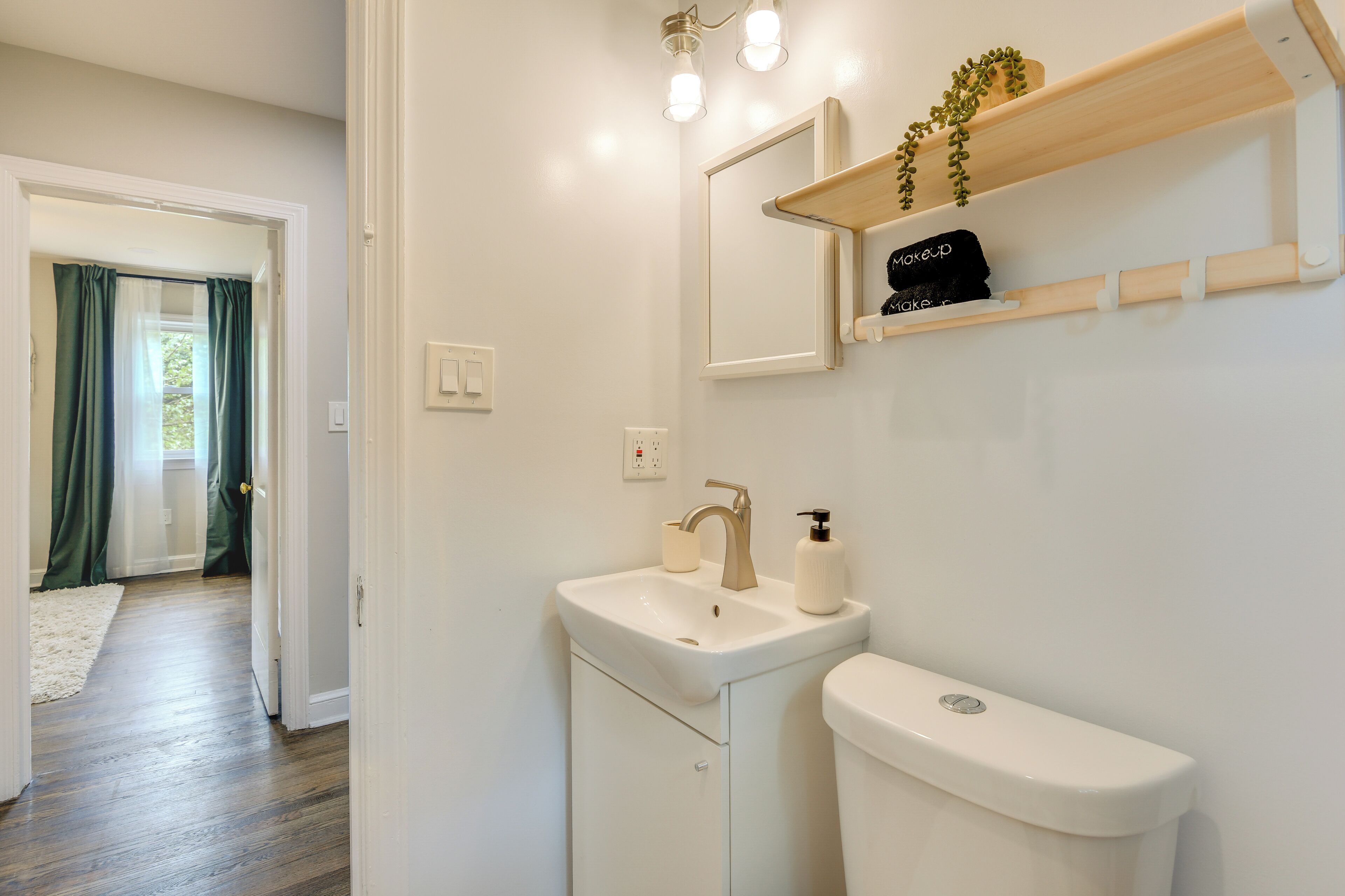 Un baño con un lavabo blanco, un espejo y un inodoro. Sobre el espejo hay un estante de madera con una planta y una bolsa de maquillaje.