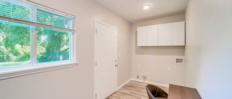 A small, well-lit room with a wooden floor, a white door, a window with blinds, and a small cabinet.