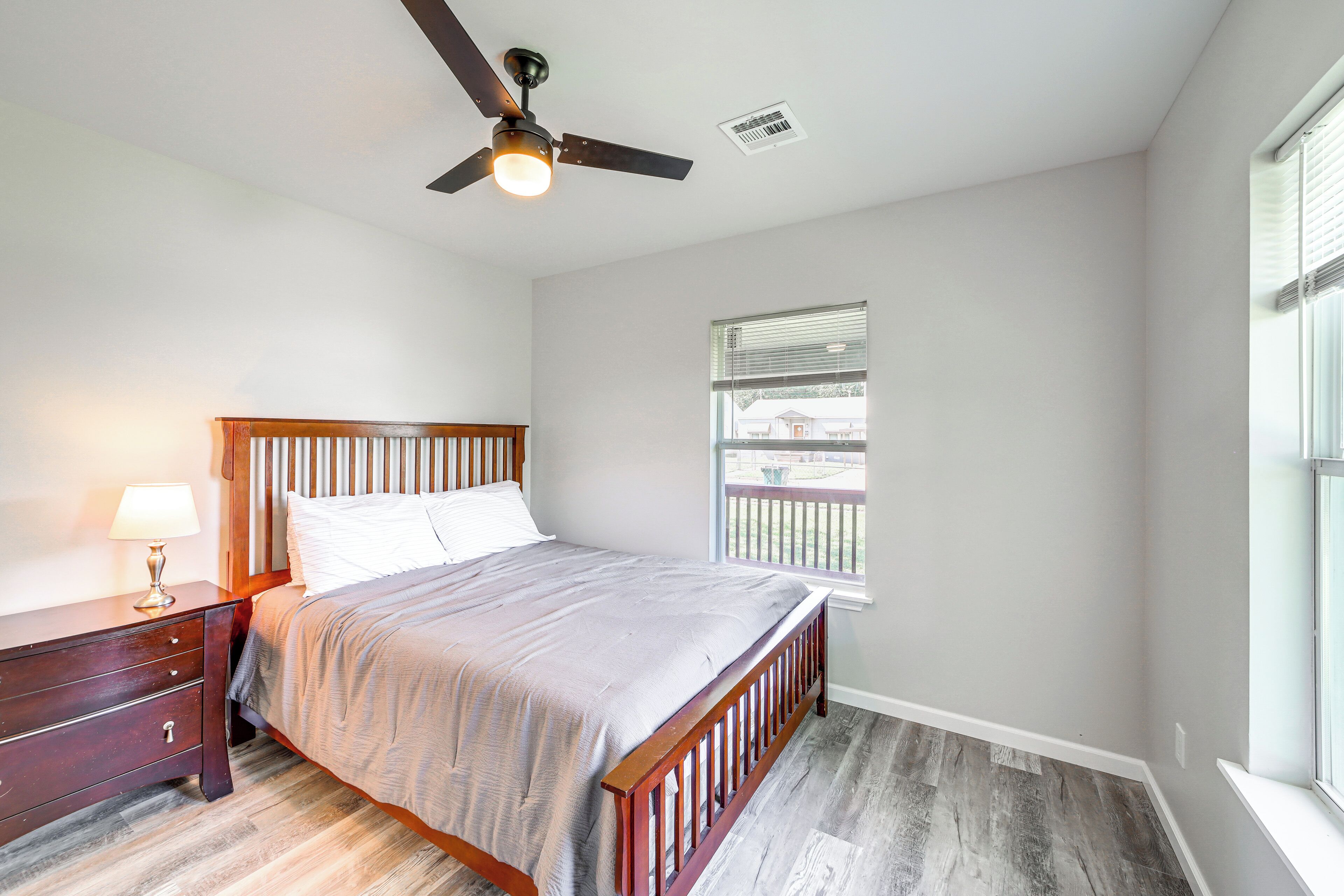 A bedroom with a bed, nightstand, lamp, ceiling fan, window, and wooden flooring.