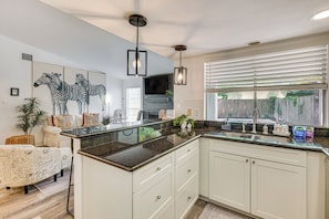 A kitchen with white cabinets, a granite countertop, and a built-in sink. There is a large window with blinds, a flat-screen TV mounted above the counter, and a zebra-themed artwork on the wall. - Pet-friendly Spring Home w/ Private Pool & Hot Tub (The Woodlands)