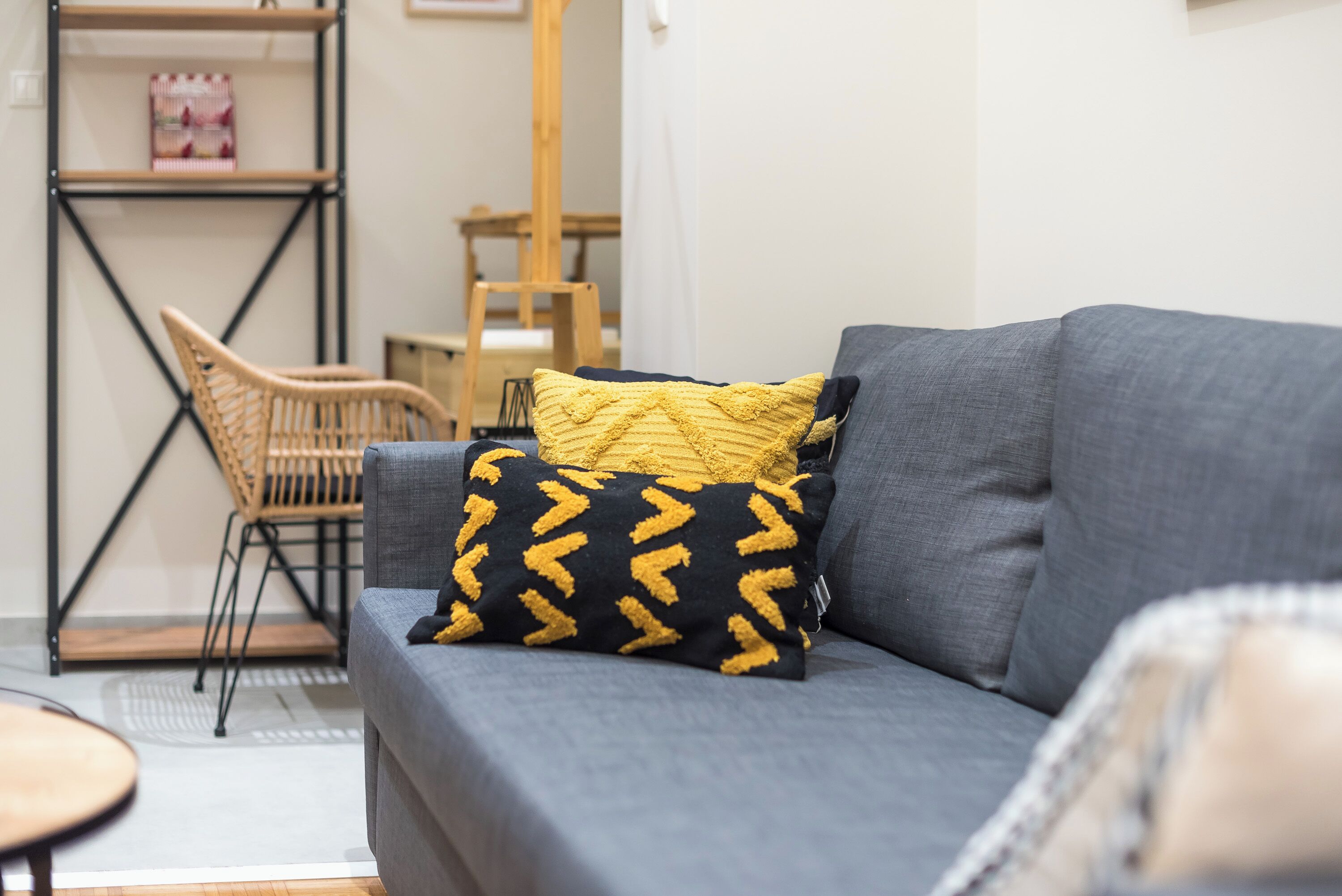 A grey sofa with two patterned cushions, a wicker chair, and a small round table in a room with a white wall and a wooden shelf.