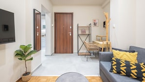 Una sala moderna con un sofá gris, cojines amarillos y negros, una puerta de madera, una planta pequeña y un televisor montado en la pared.