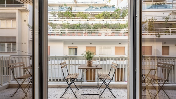 Un balcón con mesa y sillas, una planta en maceta y vistas de un edificio de varios pisos con balcones.