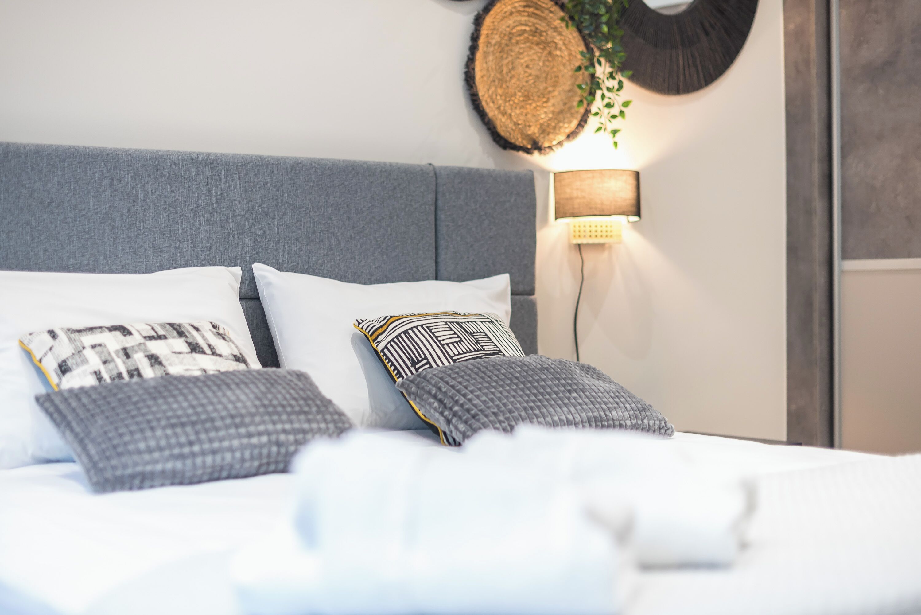 Un lit bien fait, agrémenté d’oreillers à motifs, d’une tête de lit grise et d’une lampe fixée au mur.