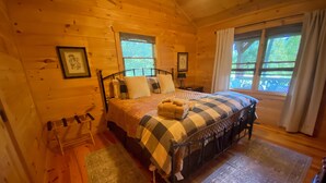 A cozy log cabin bedroom with a bed, two windows, a chair, and a rug. - Peaceful and secluded with year-round mountain views and waterfall sounds. (Cherry Log)