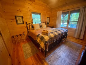 A cozy log cabin bedroom with a bed, two windows, a chair, and a rug. - Peaceful and secluded with year-round mountain views and waterfall sounds. (Cherry Log)