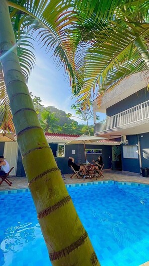 A pool area with a palm tree, a covered seating area, and a building with a balcony. - POUSADA BRISA MAR UBATUBA (Ubatuba)