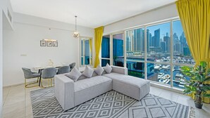 A modern living room with a large sectional sofa, a dining area with a small table and chairs, and a large window offering a view of a marina and city skyline.