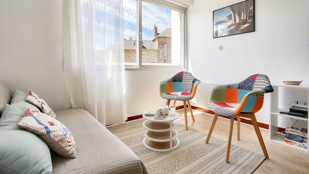 Una sala de estar con un sofá, dos sillones coloridos, un rascador para gatos y una ventana con vistas a edificios.