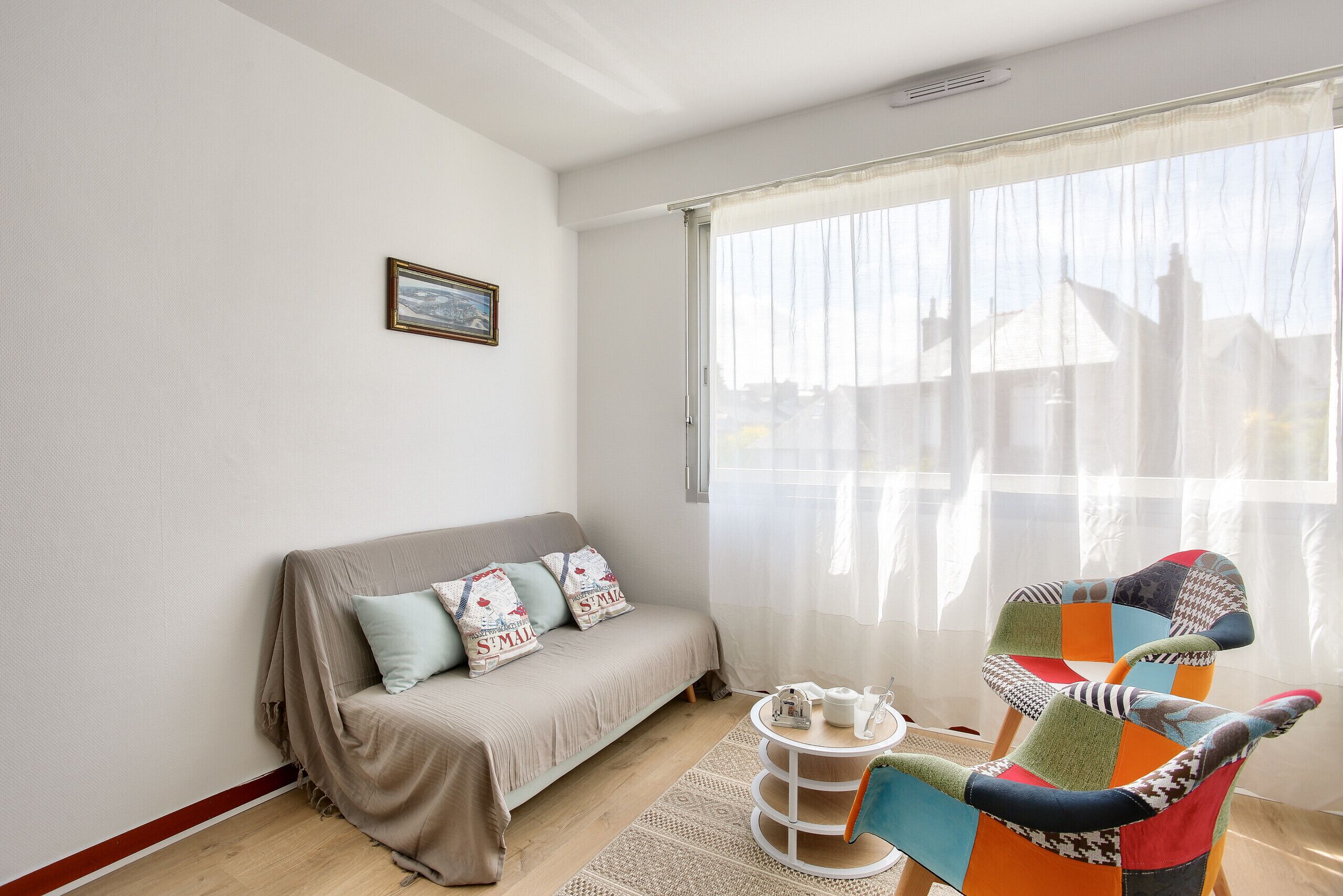 A living room with a sofa, a colorful chair, a small table with a teapot, and a window with sheer curtains.