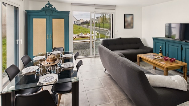 A modern living room with a glass dining table, grey sofa, and teal cabinet.