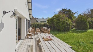 Una terraza de madera con asientos exteriores, un edificio blanco y un seto.