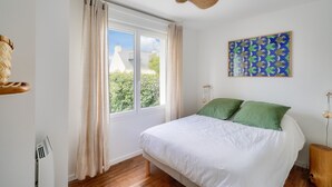 Un dormitorio con un ventanal grande, una cama con ropa de cama blanca, un cojín verde, una lámpara de techo tejida y una obra de arte colgada en la pared.