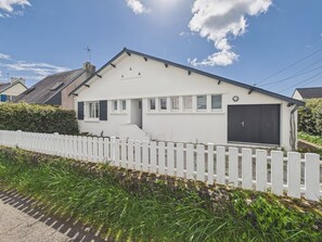 Une maison blanche à deux étages, avec une porte de garage noire, une clôture blanche en bois et un ciel bleu dégagé.