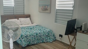A bedroom with a bed, a standing fan, a desk with a computer, and a window with blinds. - Mamita’s Beach House PR

 (Vega Baja)