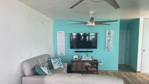 A living room with a grey sofa, teal cushions, a TV, and a ceiling fan. - Mamita’s Beach House PR

 (Vega Baja)