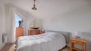 A bedroom with a large bed, a wooden nightstand, a small table with a lamp, a sailboat decoration, and a view through a window. - On the Quai de Pornic, Apartment for 4 (Pornic)