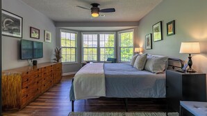 Une chambre à coucher avec un lit, un ventilateur de plafond, une télévision, une commode et de grandes fenêtres.