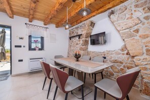 A dining area with a stone wall, a TV mounted on the wall, and a table with chairs. - Villa Melary with the Pool, Jacuzzi & Ps5 surrounded with nature. (Kras)