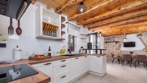 A modern kitchen with white cabinetry, a wooden countertop, and a stone wall. There is a dining area with a table and chairs. - Villa Melary with the Pool, Jacuzzi & Ps5 surrounded with nature. (Kras)