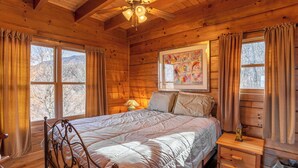 A wooden cabin bedroom with a bed, a nightstand, a ceiling fan, and a window with curtains. - Faraway Skies (Zionville)