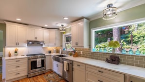 A modern kitchen with white cabinets, stainless steel appliances, a large window with a view of greenery, and a decorative vase on the counter. - Chipmunk View Lodge (Blowing Rock)