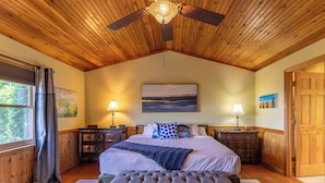 A bedroom with a large bed, wooden ceiling, two bedside tables with lamps, a bench, and a view of the outdoors through a window. - Chipmunk View Lodge (Blowing Rock)