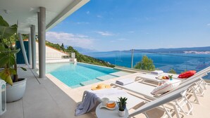 Un espace aménagé au bord de la piscine, avec des chaises longues, une table sur laquelle est posé un panier de fruits, et une vue sur la mer.