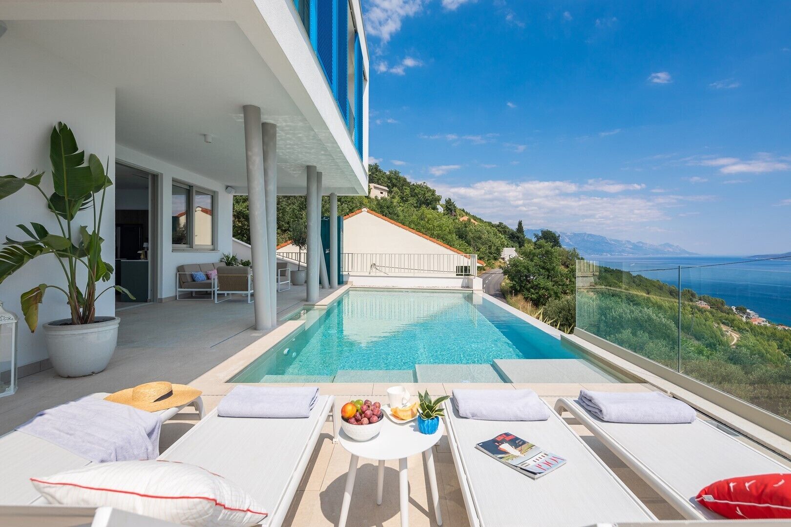 A modern outdoor pool area with lounge chairs and a table with fruit and magazines.