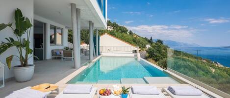 A modern outdoor pool area with lounge chairs and a table with fruit and magazines.