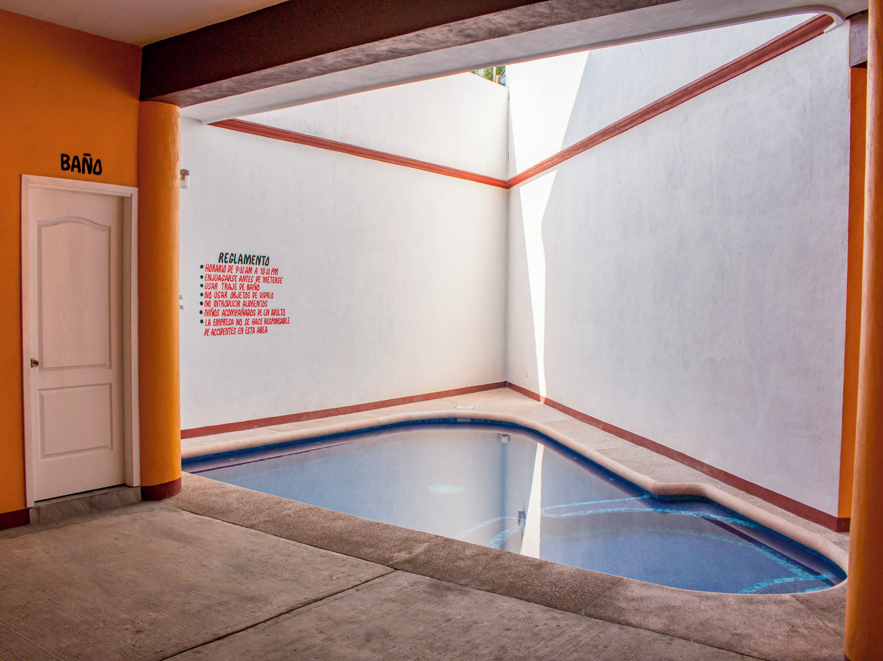 A swimming pool area with a white door labeled "BAÑO" and a sign listing rules.