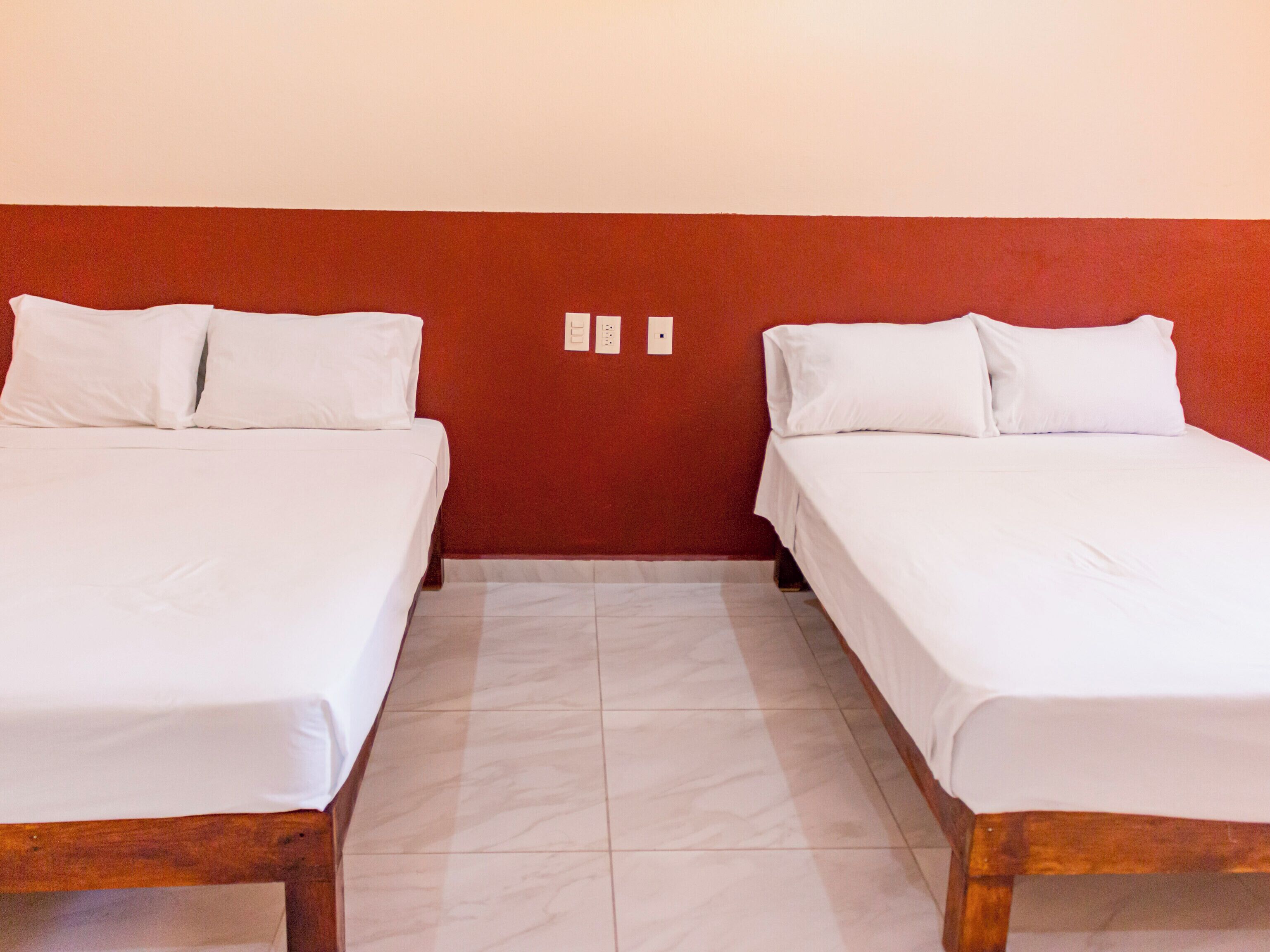A room with two single beds, each with white bedding and pillows, a wooden headboard, and a tiled floor.