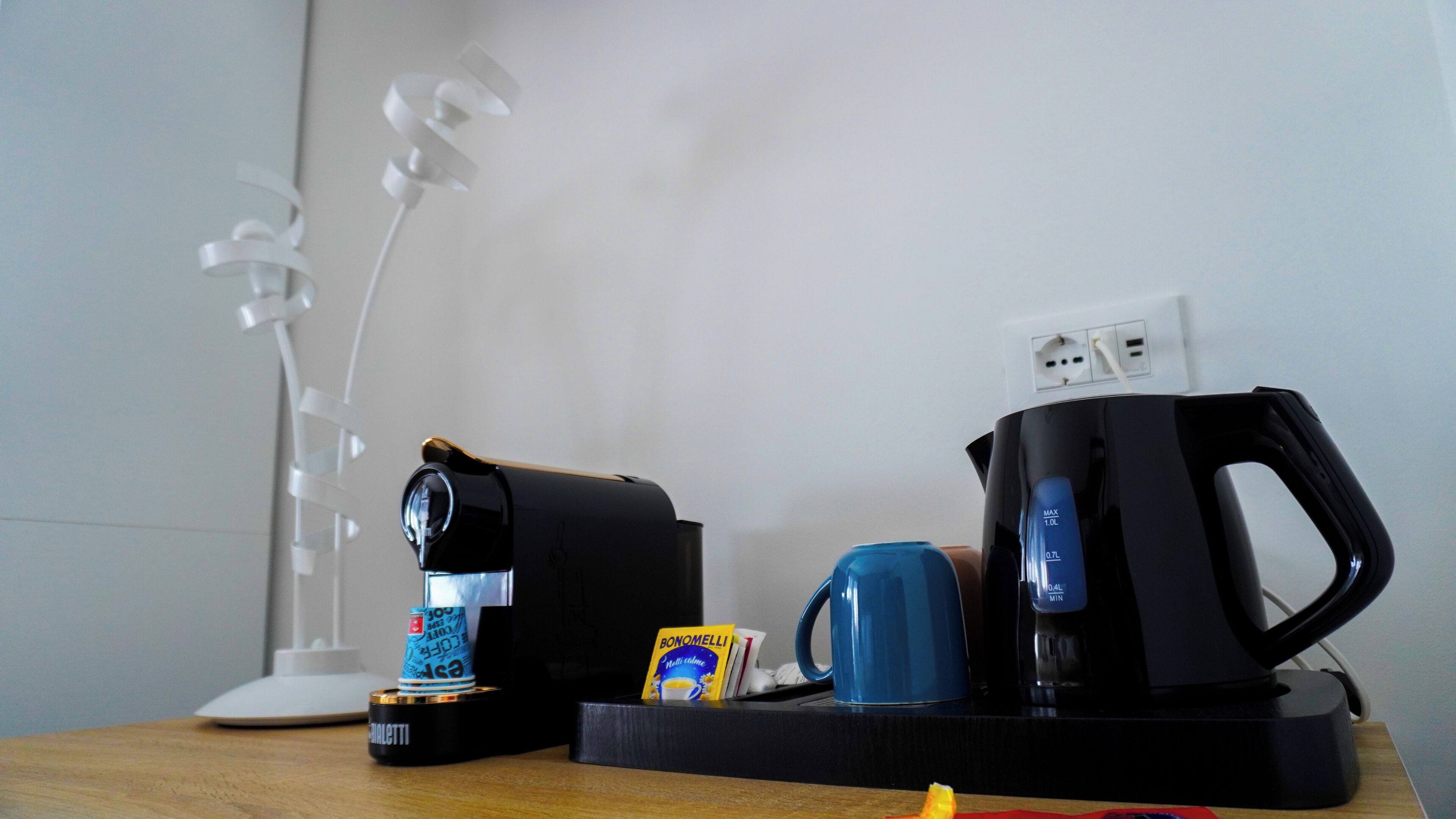 Een koffiezetapparaat, een blauwe mok, een pakje Bonomel koffie en een witte lamp op een houten tafel.
