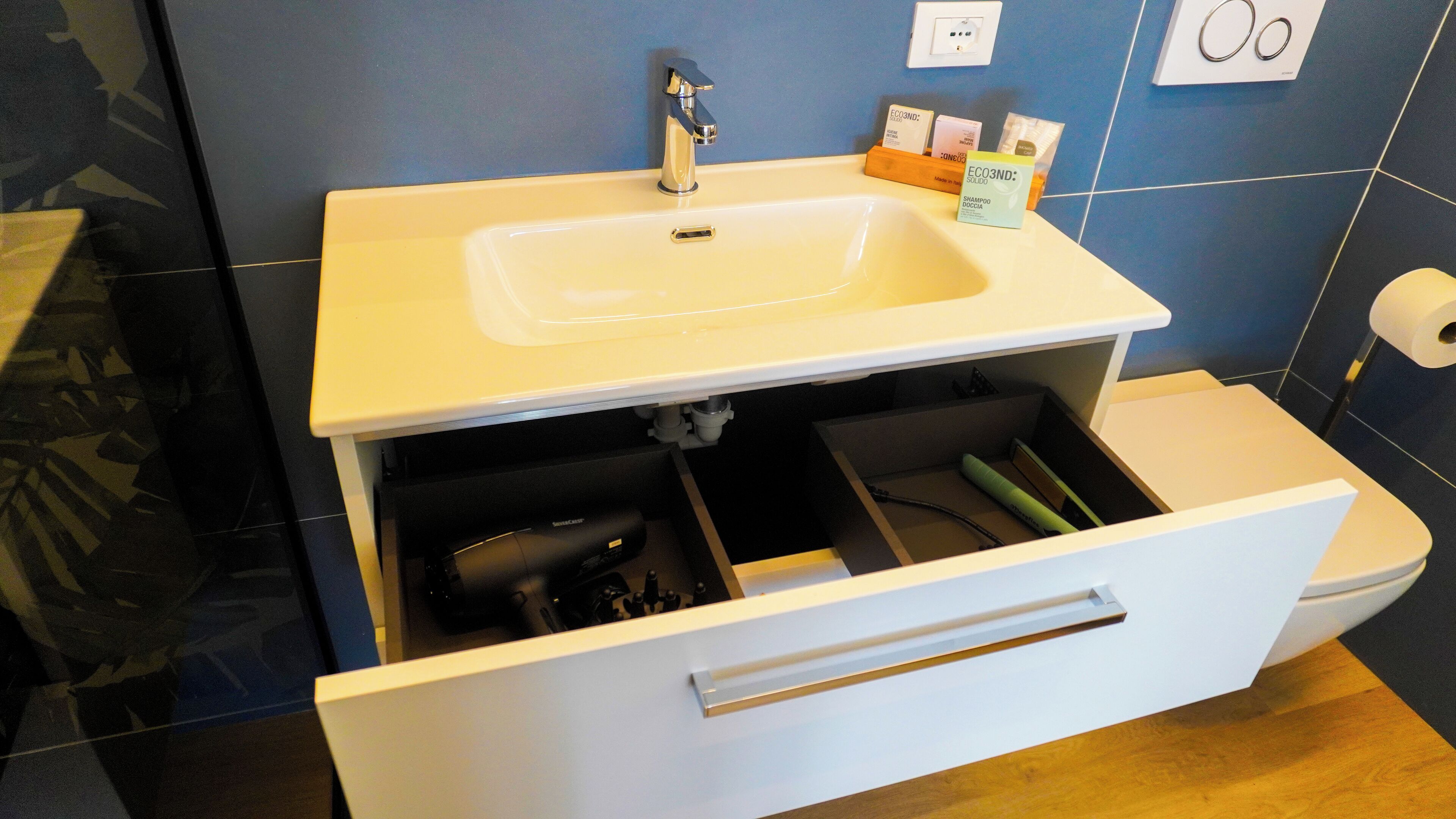 A bathroom with a white sink, a mirror, and a toilet. There is a drawer under the sink with various items.