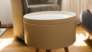 A round, light-colored ottoman with wooden legs on a wooden floor, positioned in front of a tufted armchair with a high backrest and button details, in a room with a large window and sheer curtains. - The Platform Rooms (Ferrara)